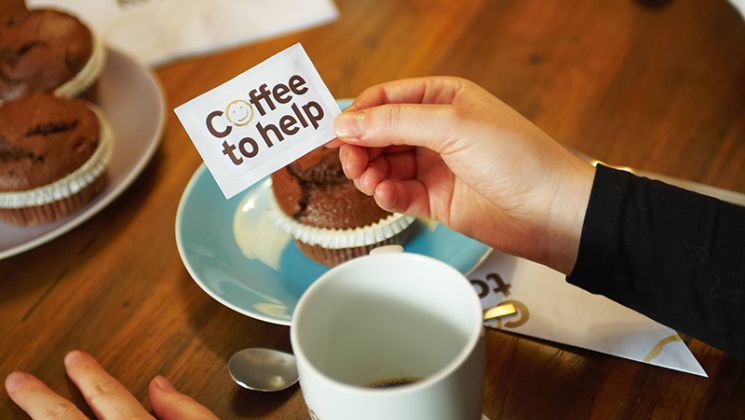Eine Hand hält ein Schild mit der Aufschrift 'Coffee to help' über einem Tisch mit Kaffee und Schokoladenmuffins.