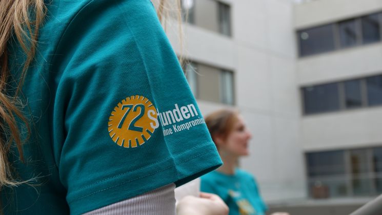 Detailansicht eines T-Shirts mit dem Logo von '72 Stunden ohne Kompromiss', in einer Outdoor-Umgebung mit Menschen im Hintergrund.