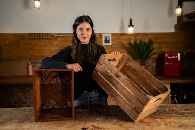 Eine Frau steht an einem Tisch und hält zwei verschiedene Holzkisten in der Hand. Der Hintergrund zeigt eine gemütliche Werkstatt.