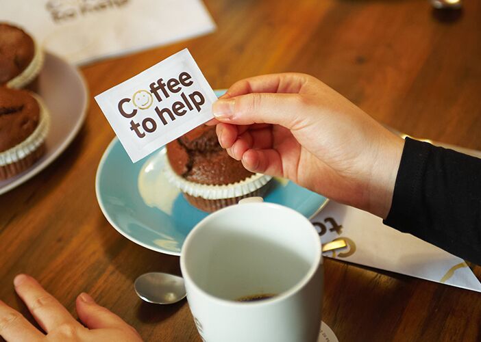 Eine Hand hält ein Schild mit der Aufschrift 'Coffee to help' über einem Tisch mit Kaffee und Schokoladenmuffins.