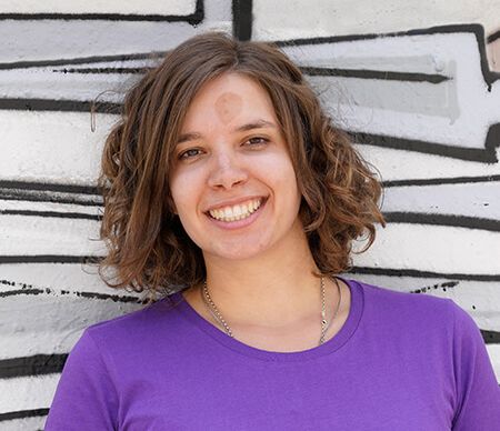 Eine junge Frau mit lockigem Haar steht vor einer grafitiartigen Wand und lächelt, trägt ein lila T-Shirt von youngCaritas.