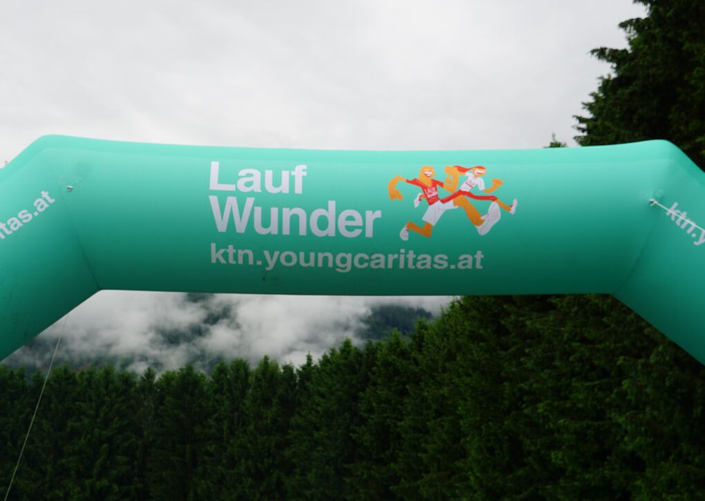 Ein leuchtend grüner Startbogen mit der Aufschrift 'Lauf Wunder' in den Bergen, umgeben von Wäldern und einem bewölkten Himmel.
