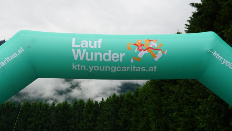 Ein leuchtend grüner Startbogen mit der Aufschrift 'Lauf Wunder' in den Bergen, umgeben von Wäldern und einem bewölkten Himmel.