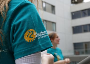 Detailansicht eines T-Shirts mit dem Logo von '72 Stunden ohne Kompromiss', in einer Outdoor-Umgebung mit Menschen im Hintergrund.